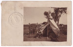 Postcard, with a photo of a man sitting outside of a tent in a rural setting (in what appears to be sparse bushland)