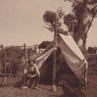 Postcard, with a photo of a man sitting outside of a tent in a rural setting